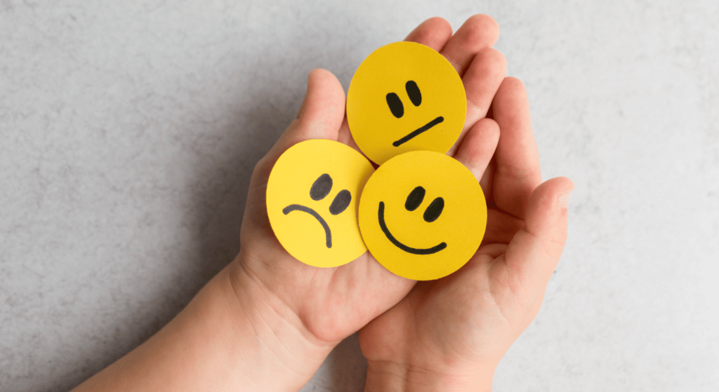 Manos sosteniendo emoticones amarillos que expresan tristeza, neutralidad y felicidad